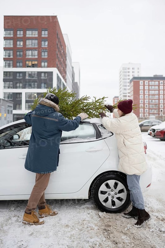 A couple wearing winter clothing secures a Christmas fir onto their car's roof in a snow-covered city setting Buildings in the background provide an urban holiday atmosphere