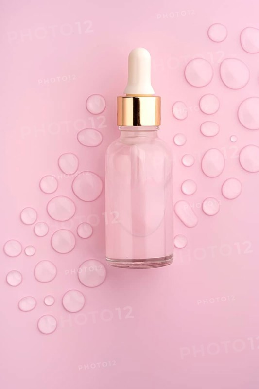 A clear glass dropper bottle on a pink background, surrounded by scattered water drops The scene evokes freshness, beauty, and skincare elegance
