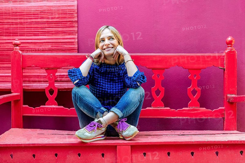 A woman wearing a blue plaid shirt sits cross-legged on red wooden stairs, smiling gently and looking away against a vibrant pink wall