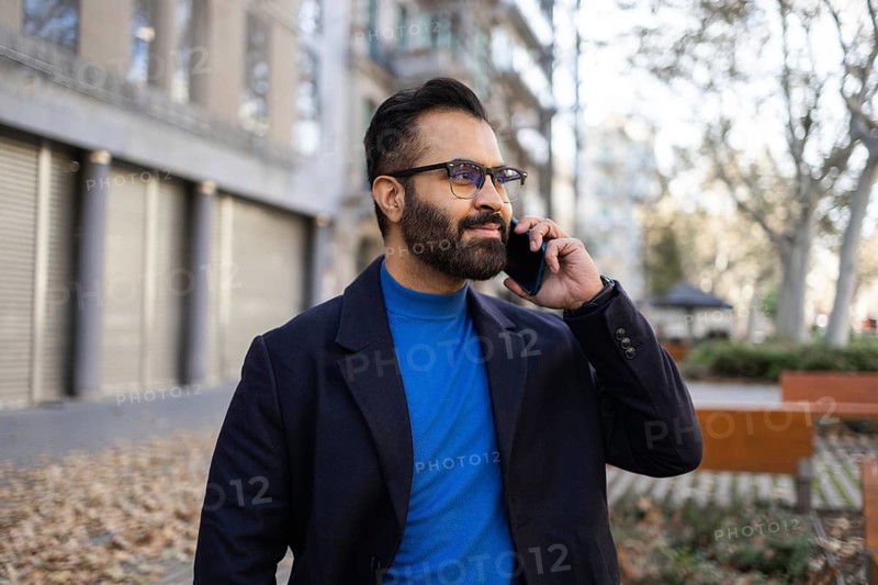 Indian businessman in winter clothing using a mobile phone in an urban park setting, looking away from the camera