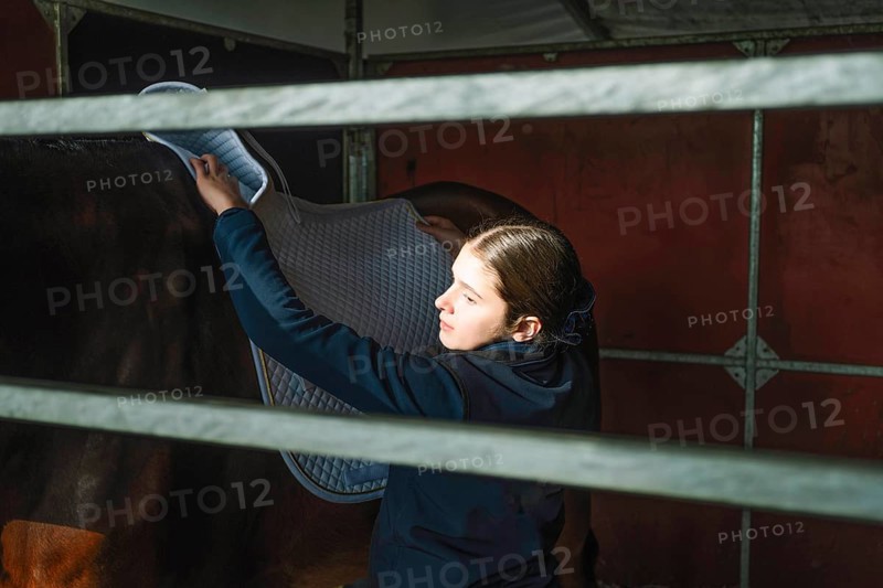 A teenage girl carefully places a saddle blanket on a horse in preparation for a classical dressage session, demonstrating focus and care inside a stable