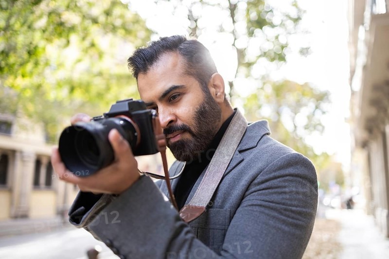 An Indian man, dressed professionally in a grey coat, focuses intently on capturing a photo with his advanced camera on a sunny city street
