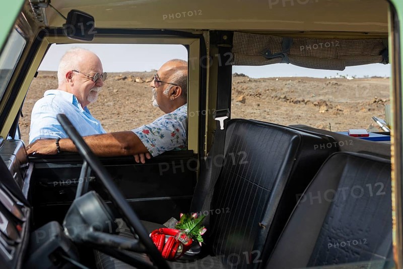 A senior gay couple shares a joyful moment inside an off-road vehicle, engaging in conversation with clear affection. Both men, likely in their late sixties or early seventies, are casually dressed and appear to be relishing their outdoor adventure