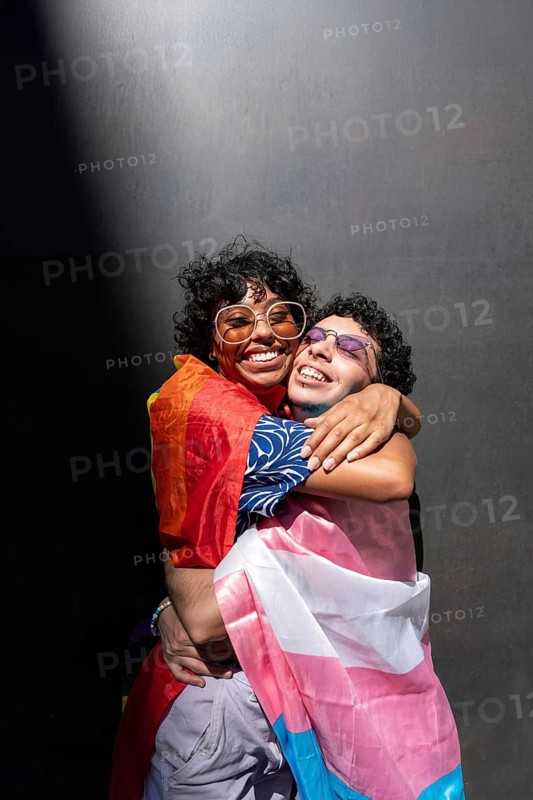 A joyful, multiethnic LGBTQ+ couple embraces warmly, each draped in vibrant pride flags, symbolizing love and diversity against a dark background