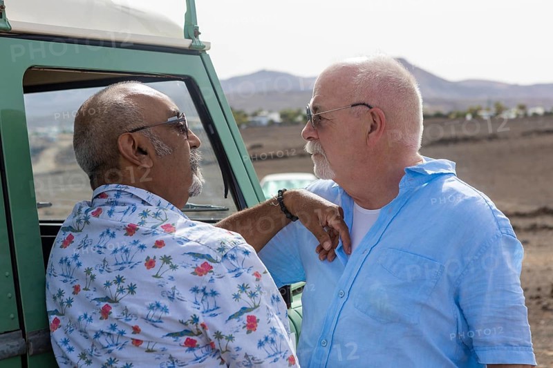 A senior gay couple shares a tender moment beside their off-road vehicle during a camping trip. Both men gaze at each other lovingly, emphasizing a warm, affectionate connection in a serene natural setting