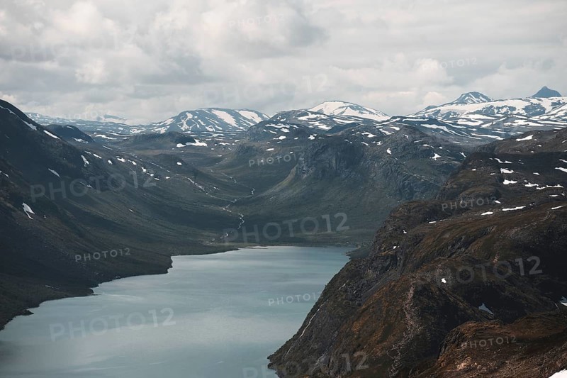 A sweeping view of a tranquil lake nestled in a dramatic mountain valley in Norway Snow-dusted peaks and cloudy skies frame this breathtaking Nordic wilderness scene