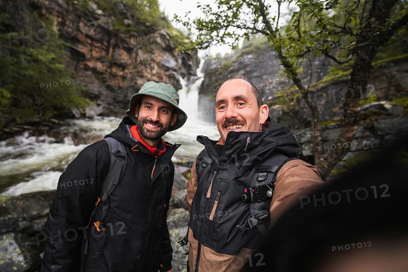 Two adventurers enjoying a scenic road trip in Norway Surrounded by lush forests and a rushing waterfall, they capture the essence of trekking and camping in the wilderness