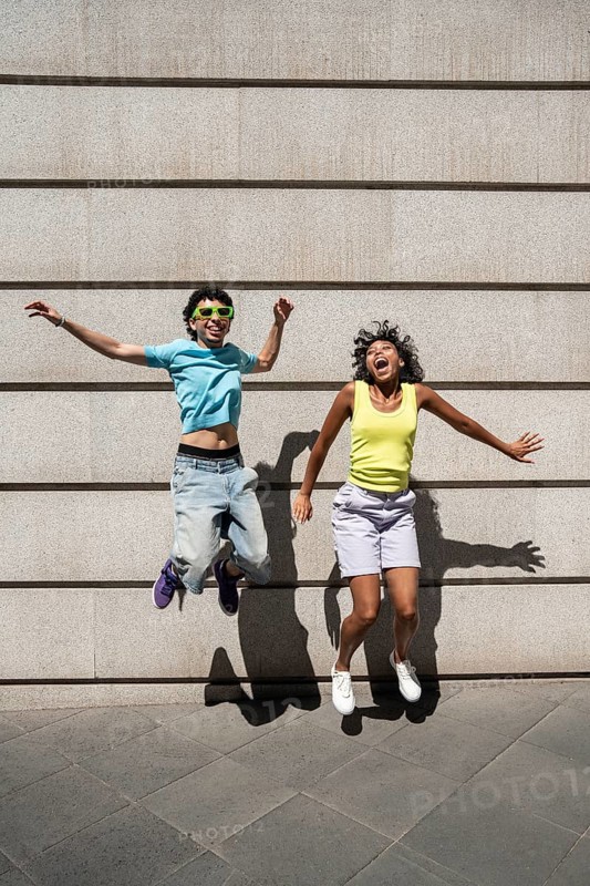 A joyful, multiethnic LGBTQ+ couple, wearing vibrant summer attire, jumps against a concrete wall, symbolizing happiness and diversity on a sunny day