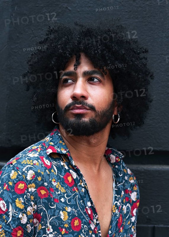 Portrait of a stylish man with curly hair wearing hoop earrings and a vibrant floral shirt. The image captures modern fashion and urban style against a gray background