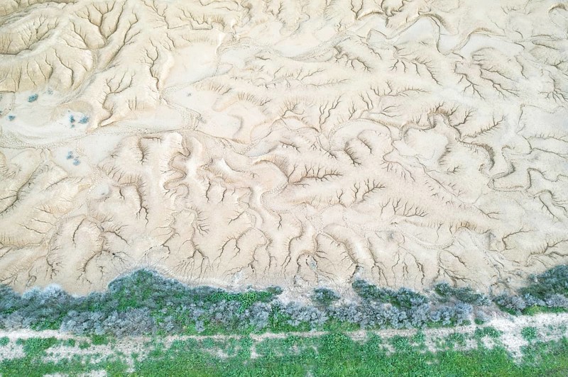 Aerial view of a uniquely textured, arid landscape showcasing patterns of erosion A strip of green vegetation lines the bottom, contrasting the dry terrain above