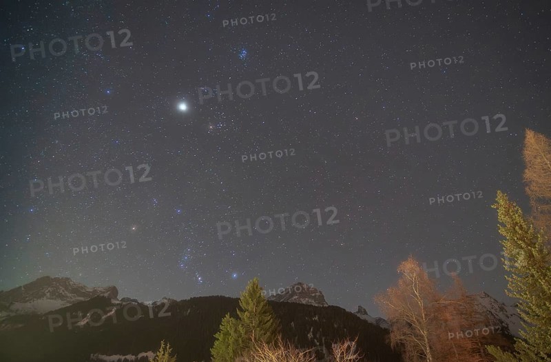 The constellations of Orion and Taurus shine brightly in the night sky, accompanied by Jupiter, Sirius, the brightest star, and the Pleiades. Snowy alpine peaks and trees enhance the celestial view