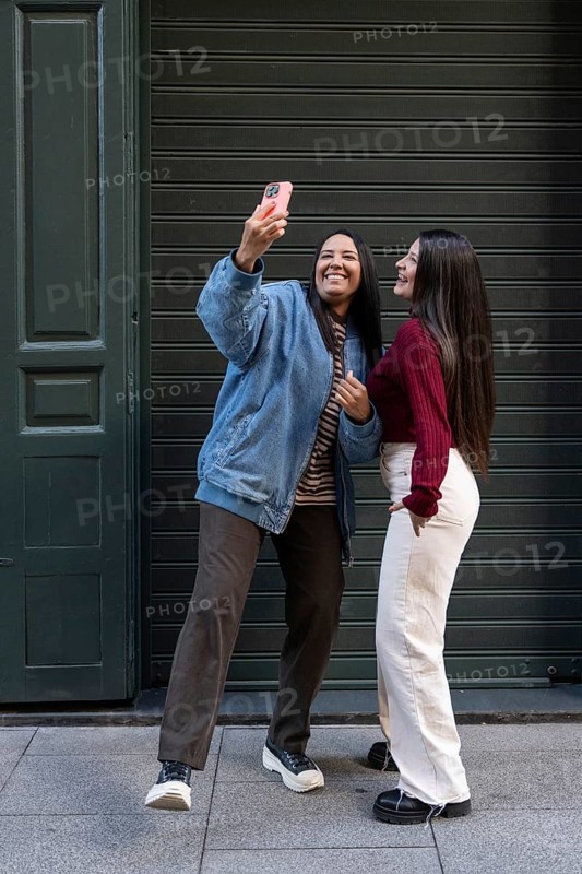 A joyful latin lesbian couple poses for a selfie in an urban setting, showcasing their unity and happiness. The relaxed atmosphere conveys warmth and a strong connection between them