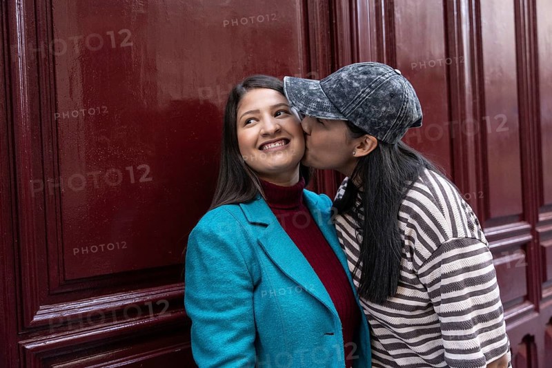 A loving Latin lesbian couple shares a sweet moment by a burgundy wall. One partner playfully kisses the other on the cheek, radiating affection and happiness