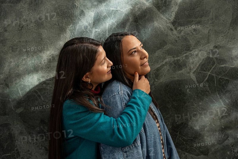 A Latin lesbian couple shares a loving embrace, expressing warmth and affection. The tender moment is captured against a textured background, highlighting their connection