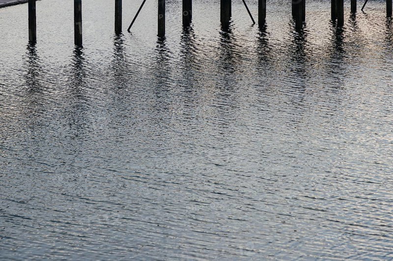 Water reflections with wooden poles standing vertically. The ripples and light reflections create a calm and textured scene