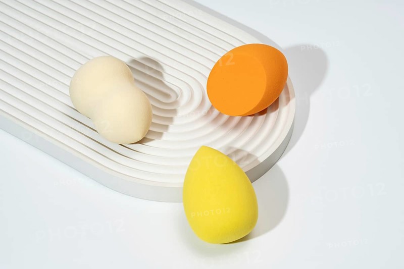 A trio of colorful cosmetic sponges in orange, yellow, and cream are arranged on a textured, white surface The minimalist composition highlights the beauty tools in soft, natural lighting