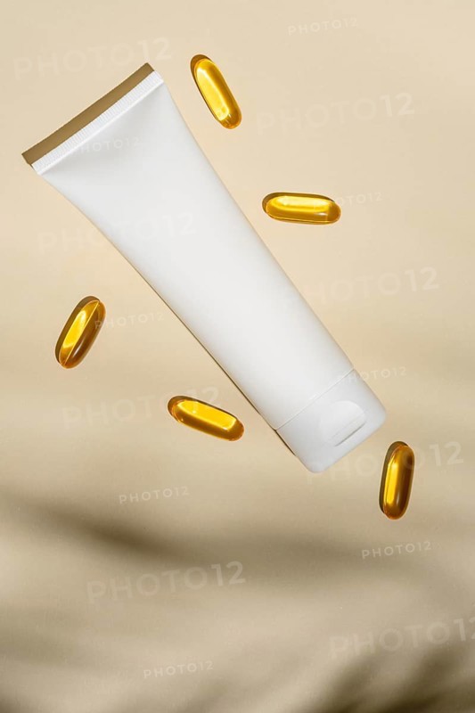 Top view of a blank white cosmetic tube floats amidst golden oil capsules, all set against a soft beige background, suggesting luxury skincare