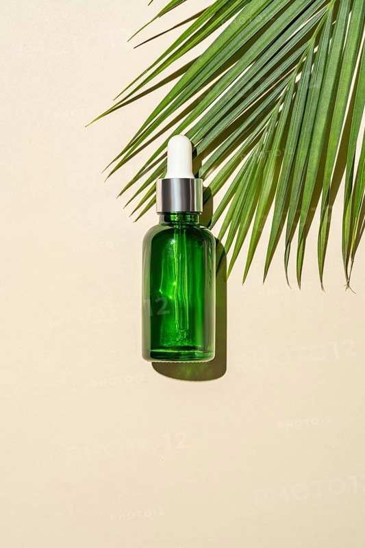 Top view of a green glass serum bottle with a dropper pipette, cast by a palm leaf's on a warm beige background, symbolizing organic skincare