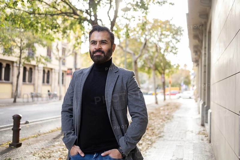 An Indian man, dressed as a businessman in winter attire, strolls along a city street with his hands in pockets, looking away