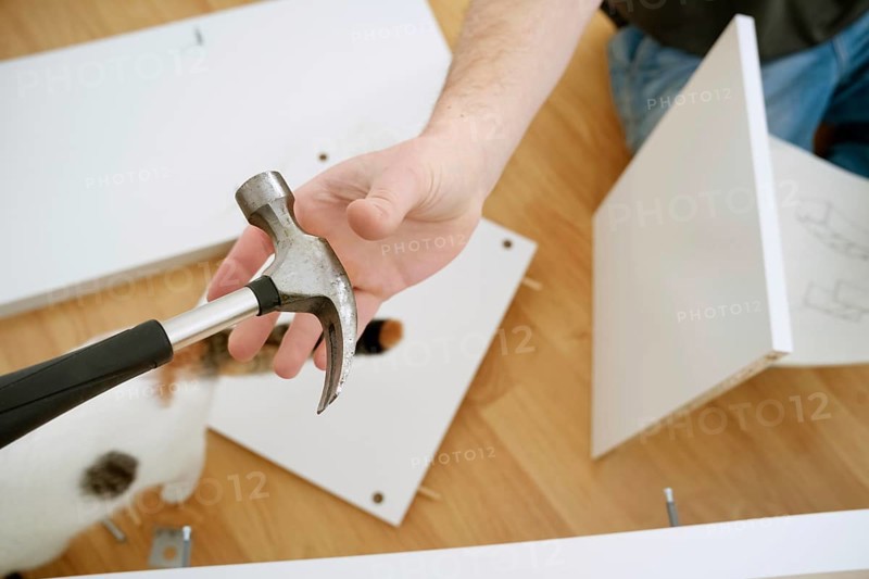 Cropped view of a man holding a hammer while assembling new furniture. The image shows his hands, the tool, and parts of the furniture on a wooden floor