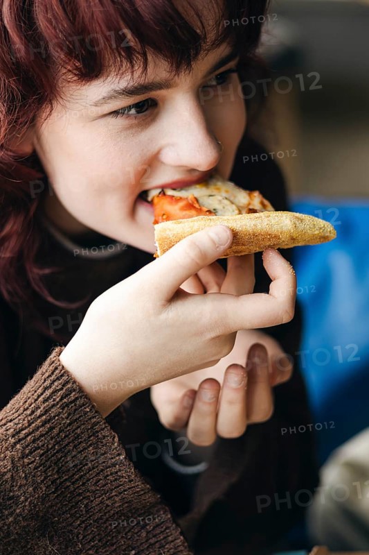 A young woman with red hair joyfully eats a slice of pizza, focusing on the flavor and her dining experience