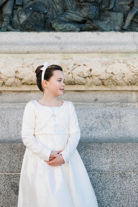 A young girl stands in a beautiful First Communion dress, framed by an ornate stone wall with intricate carvings, capturing the essence of a cherished religious milestone
