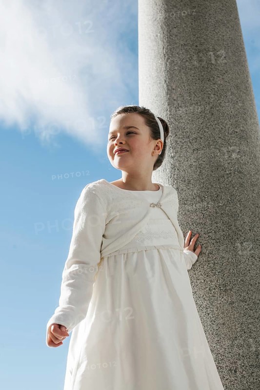 A young girl wearing a white First Communion dress stands outdoors against a stone column. The clear blue sky and clouds create a serene and joyous atmosphere