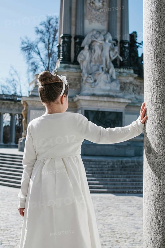 A young girl wearing a white First Communion dress stands beside a stone column, with a historic monument in the background, under a clear blue sky