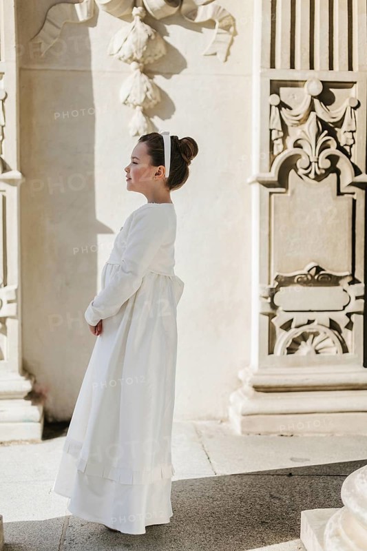A young girl wearing a First Communion dress stands by an ornate wall. The scene captures a serene moment, highlighting her elegance and the architectural beauty around her