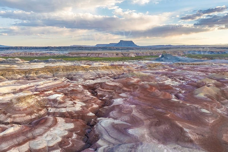 Stunning aerial photograph captures the unique patterns and vibrant colors of bentonite hills in Utah. The landscape showcases intricate natural formations set against a backdrop of vast skies and distant plateaus