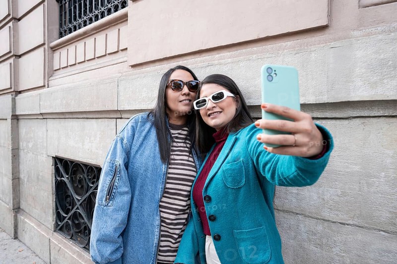 A Latin lesbian couple takes a joyful selfie outdoors, leaning against a historic building. Both wear stylish jackets and sunglasses, capturing a moment of love and happiness
