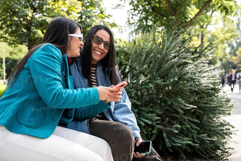 A joyful Latin lesbian couple sits on a park bench, sharing a moment with their smartphones under the sun. They smile and engage deeply, surrounded by lush greenery