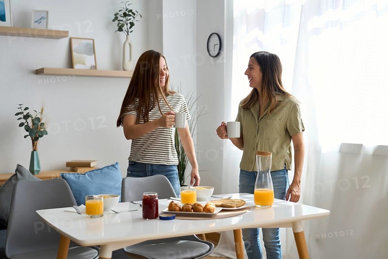 A lesbian couple shares a cozy morning breakfast at home They smile warmly at each other, holding coffee mugs, surrounded by a bright and inviting atmosphere