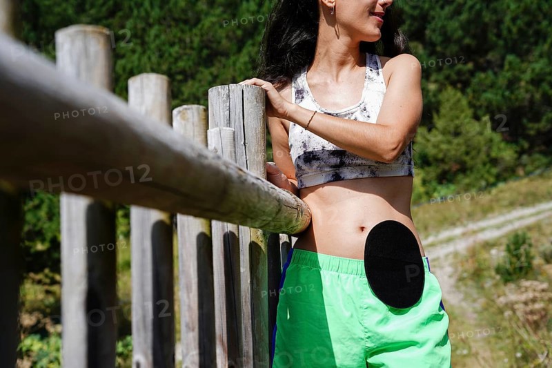 A woman with a visible ostomy bag stands confidently in a serene outdoor setting She wears a sports bra and shorts, embodying strength and embracing life