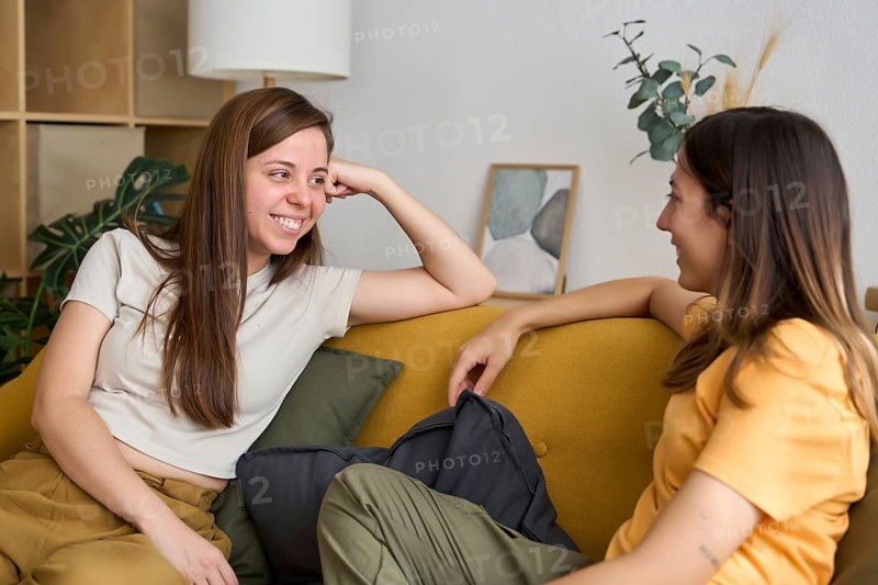 A lesbian couple shares a cozy moment on the sofa, exchanging smiles and enjoying each other's company in a warm, intimate living room setting Perfect representation of love