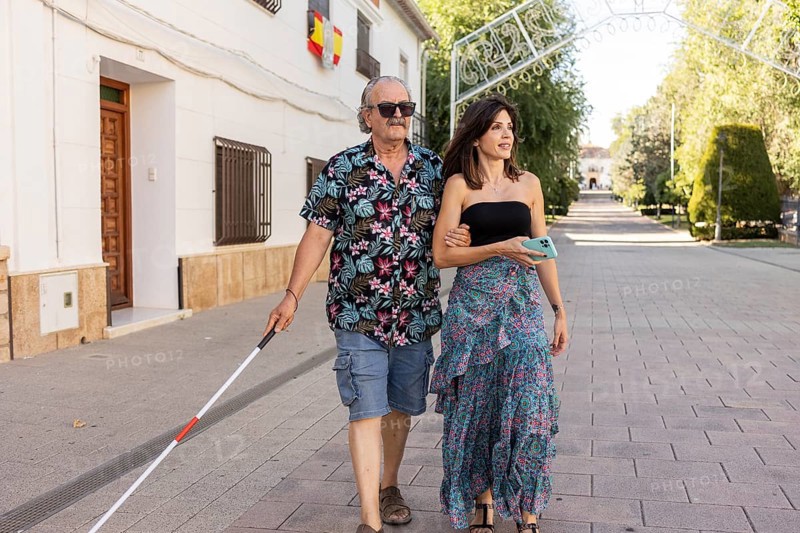 A blind father, utilizing a white cane, walks confidently through a town, guided by his attentive daughter on a sunny day
