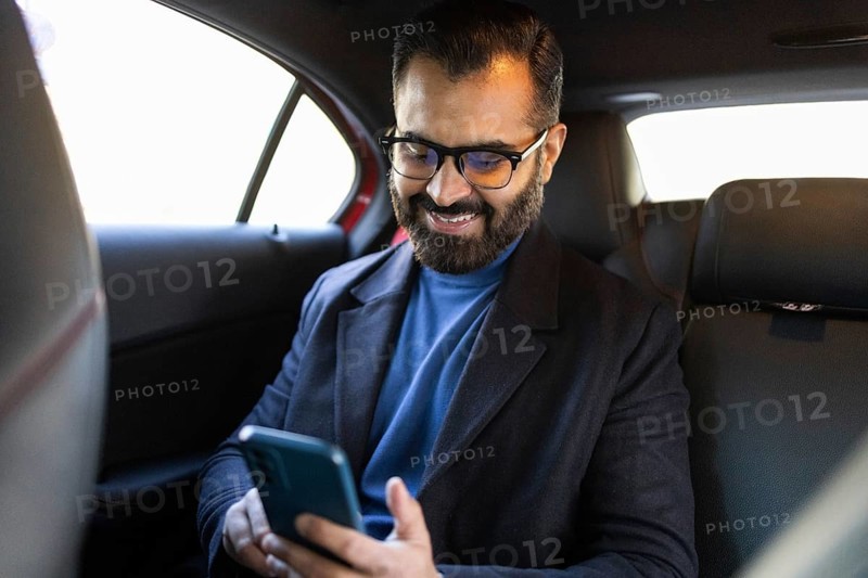 A happy Indian man, looking down at his smartphone, sits in a car, dressed in a winter coat and eyeglasses