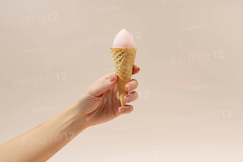 A hand holds a waffle cone with a pink cosmetic sponge inside, resembling ice cream The neutral background enhances the creative contrast of this beauty-themed image