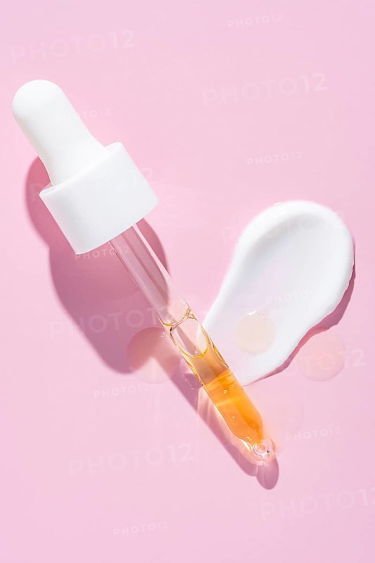 A dropper dispenses golden serum beside a white cream smear on a vibrant pink surface, representing skincare textures