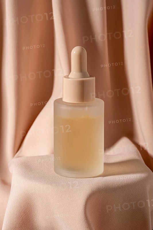 A minimalist frosted glass bottle with a dropper, containing skincare serum, is placed against luxurious soft fabric The warm beige tones create an elegant, calming ambiance