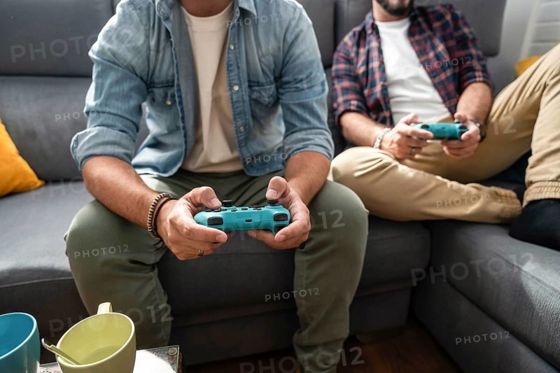 A gay couple sits on a couch, immersed in a video game. They both hold controllers, wearing casual clothes, with mugs nearby on a table, illustrating a cozy, relaxed atmosphere