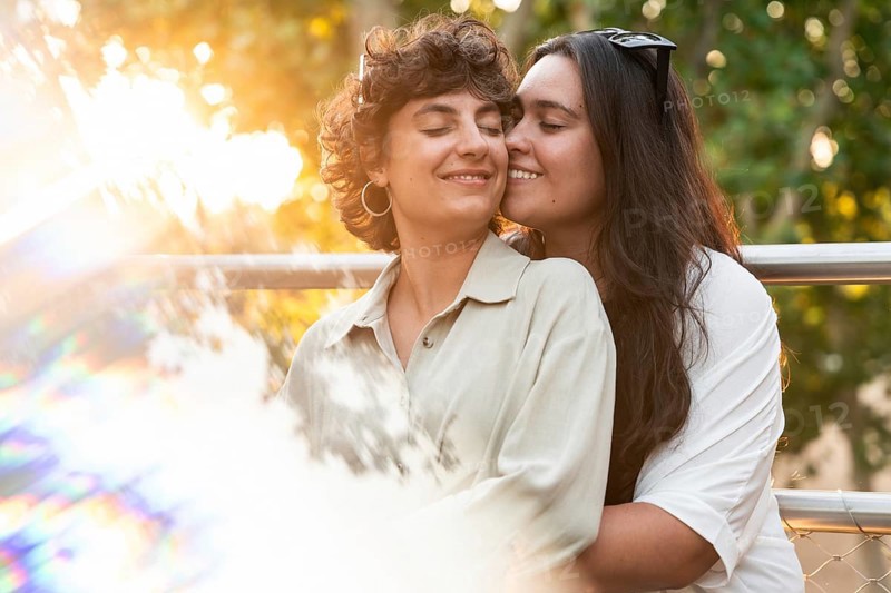 A loving lesbian couple shares a tender embrace outdoors, surrounded by vibrant nature. The sunlight adds a warm and serene ambiance to their affectionate moment