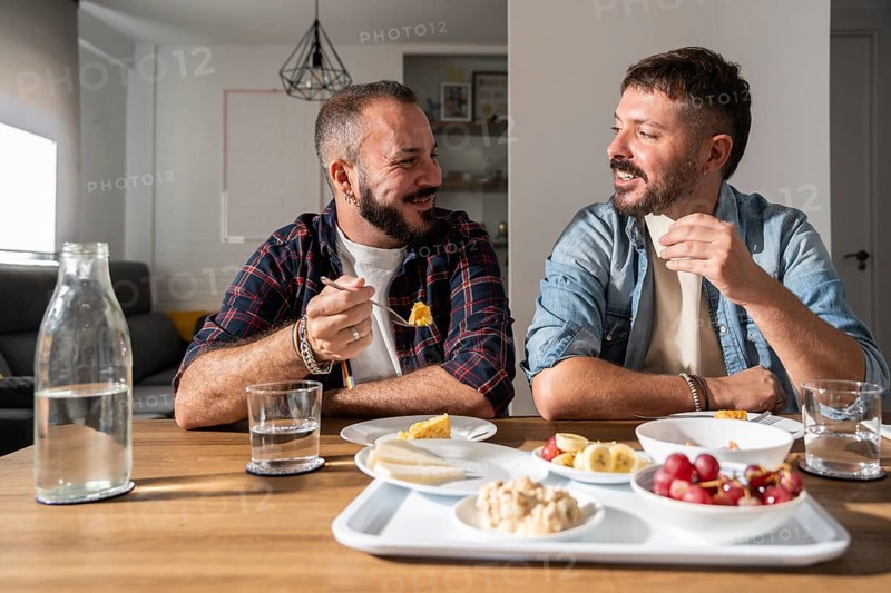 A happy gay couple sits together in a bright dining room, sharing a meal and enjoying each other's company in a relaxed home environment