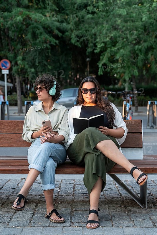 Lesbian couple relaxes on a park bench. One is reading a book, while the other listens to music, phone in hand. Surrounded by greenery and enjoying the tranquil setting