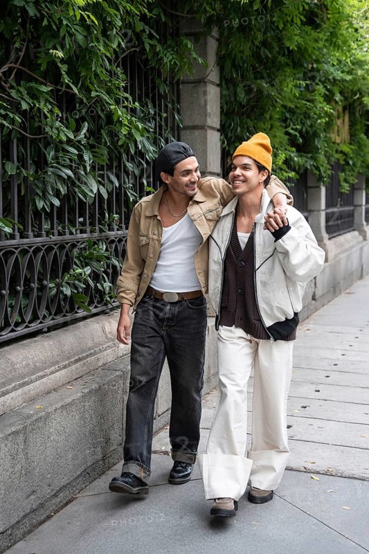 A joyful gay couple strolls arm in arm along a leafy city sidewalk, sharing smiles and enjoying each other's company on a bright day, depicting a loving relationship