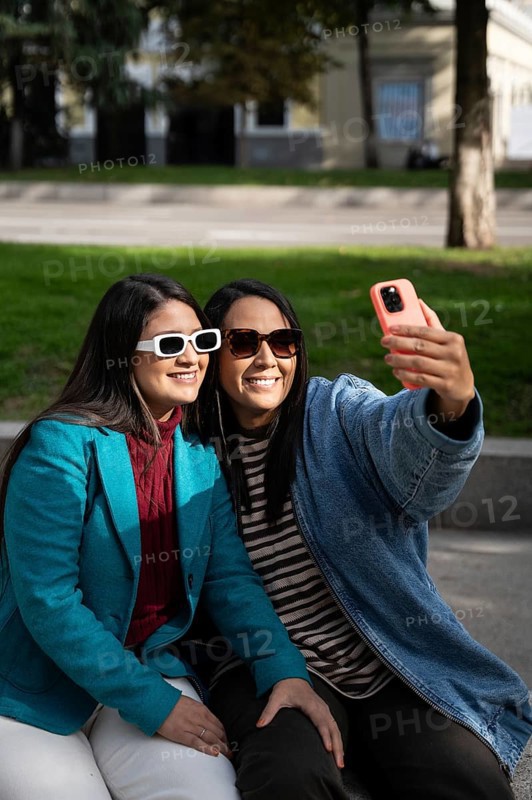 A smiling Latin lesbian couple enjoys a sunny day at the park, embracing while taking a selfie with their smartphone, capturing a joyful moment together