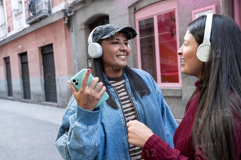A joyful Latin lesbian couple wearing headphones and smiling while listening to music. They are standing on an urban street, enjoying their time together and sharing music