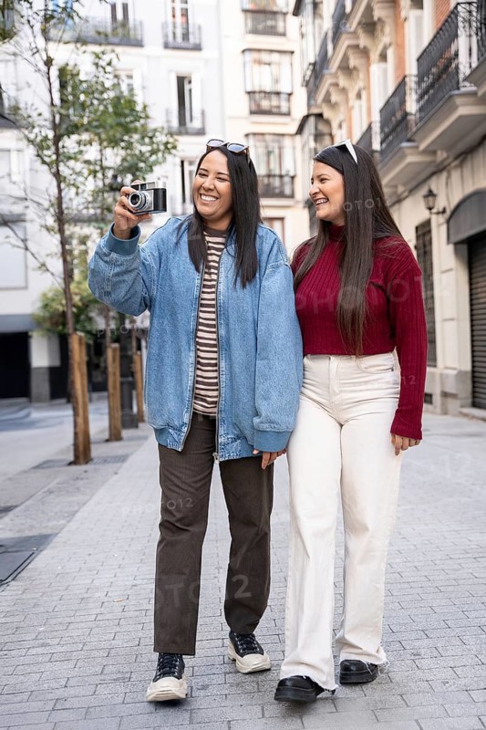 A joyful latin lesbian couple enjoys a sunny stroll, capturing memories on a city street. They share laughter, embodying love and happiness in an urban setting, creating beautiful moments together