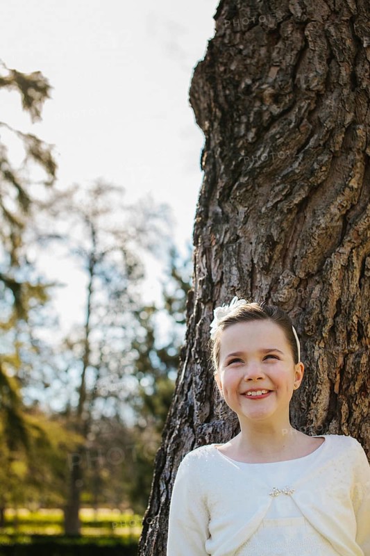 A joyful girl in a white First Communion dress stands beside a tree, beaming with happiness. Sunlight filters through the branches, creating a serene outdoor scene