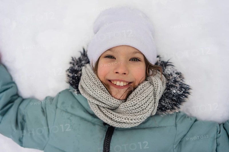 A smiling child lies in the snow, wearing a warm jacket, white hat, and knitted scarf. The joy and warmth of a winter day are captured in this delightful image